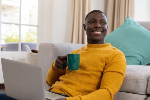 自宅のコーヒーを飲みながらソファに座ってノートパソコンを使ってアフリカ系アメリカ人の笑顔 包括性 国内生活 遠隔作業 — ストック写真