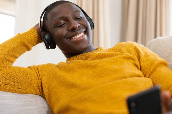 坐在沙发上 头戴耳机 看着智能手机 微笑着坐在家里的非洲裔美国人 家庭生活 闲暇时间和放松 独自呆在家里 — 图库照片