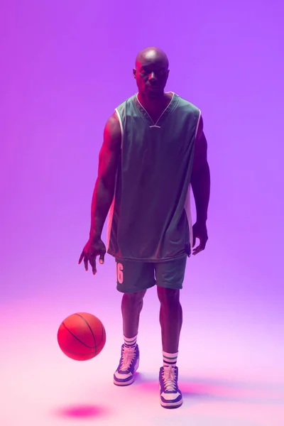 Image of african american basketball player bouncing basketball on neon purple background. Sports and competition concept.