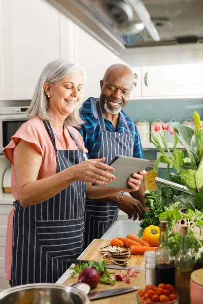 エプロンを着用し キッチンでタブレットを使用して幸せな高齢者の多様なカップル 家庭や退職のコンセプトで質の高い時間を過ごす — ストック写真
