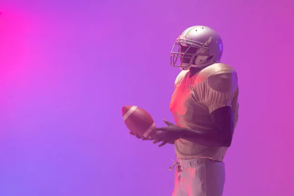 Afroamerykanin Męski Amerykański Piłkarz Trzyma Piłkę Neonowym Różowym Fioletowym Oświetleniem — Zdjęcie stockowe