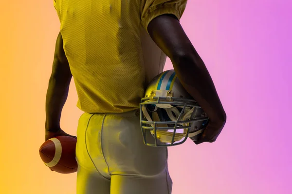 Midsection of african american male american football player with neon purple and yellow lighting. Sport, movement, training and active lifestyle concept.