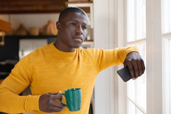 キッチンの窓から外を見てコーヒーとスマートフォンを持っているアフリカ系アメリカ人の男を考えた 包括的で国内生活家で一人で過ごす — ストック写真
