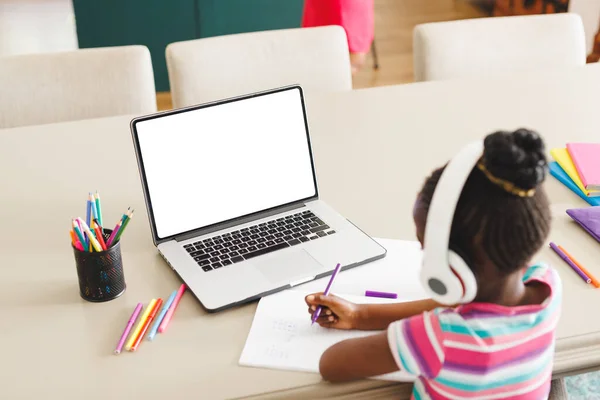 在笔记本电脑上学习时戴耳机写笔记的非洲裔美国女孩的高视角 复制空间 电子学习 无线技术 — 图库照片