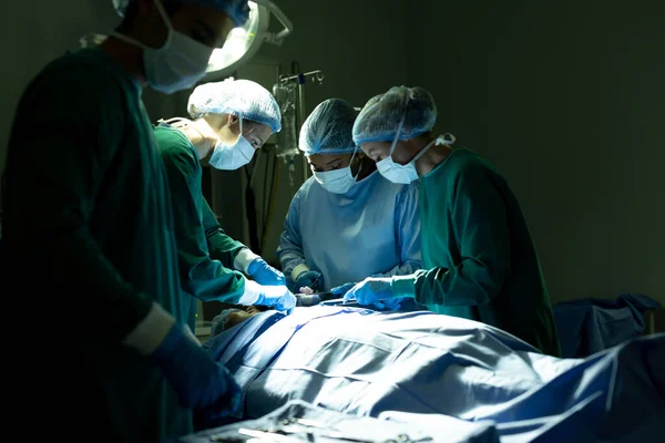 在黑暗手术室里为病人做手术的女医生和男性技术专家组成了一个多样化的小组 医疗和保健服务 — 图库照片
