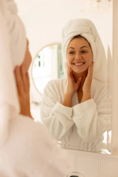 快乐的高加索女人穿着长袍 在镜子里看着自己的倒影 在浴室里微笑 独自在家度过美好时光 — 图库照片