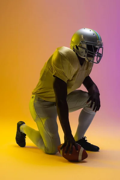 African american male american football player holding ball with neon yellow and purple lighting. Sport, movement, training and active lifestyle concept.