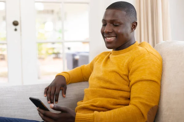 アフリカ系アメリカ人の男性が家でスマホを使ってソファに座って笑っている 包括性 国内生活 自由な時間とリラクゼーション 自宅で一人で時間を過ごす — ストック写真