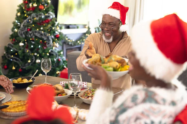 幸せなアフリカ系アメリカ人の家族は サンタの帽子をかぶって テーブルに座って夕食を食べる クリスマスに家で家族の時間を過ごす — ストック写真