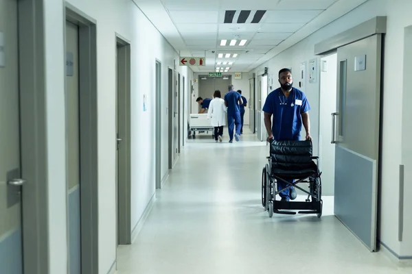 Biracial male healthcare worker pushing empty wheelchair in busy hospital corridor. Hospital, medical and healthcare services.