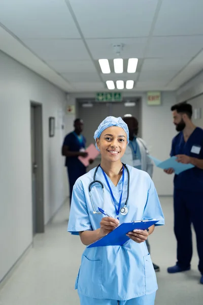 Vertical Smiling Biracial Female Doctor Writing Notes Busy Hospital Corridor — Foto de Stock