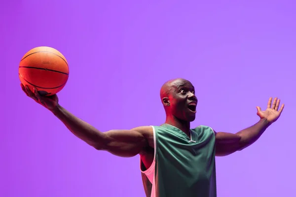 Image of smiling african american basketball player with basketball on neon purple background. Sports and competition concept.