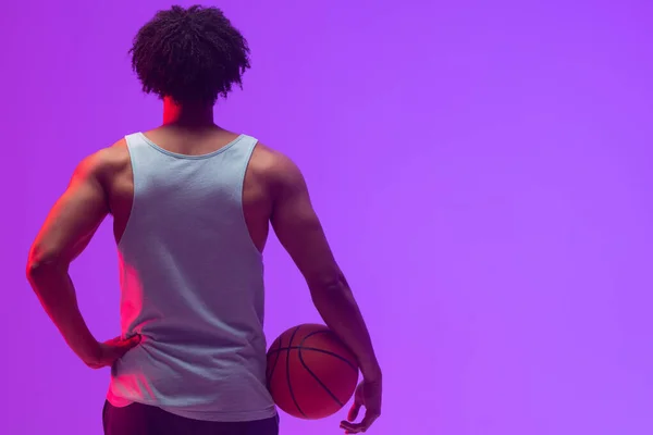 Image of rear view of biracial basketball player with basketball on neon purple background. Sports and competition concept.
