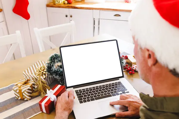 サンタの帽子をかぶった白人男性がキッチンのテーブルに座り コピースペース付きのノートパソコンを使用しています クリスマスだけで家で質の高い時間を過ごす — ストック写真