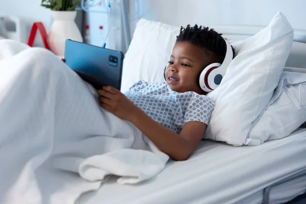 快乐的非洲男孩病人在耳机使用平板在医院的床上 医疗和保健服务 — 图库照片