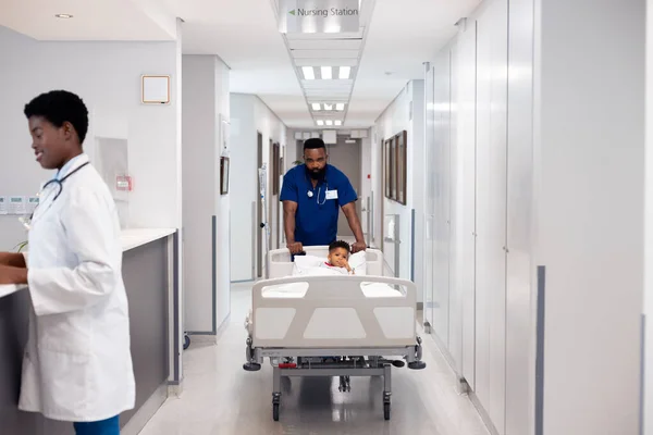 コピースペースの忙しい廊下で病院のベッドで少年患者を押すアフリカ系アメリカ人男性医師 医療サービス — ストック写真