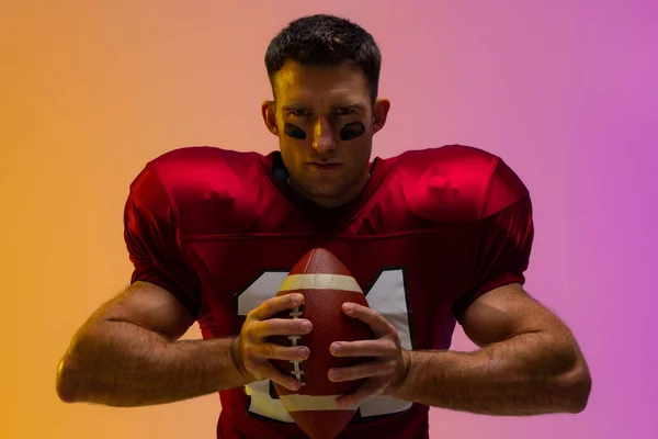 Caucasian male american football player holding ball with neon yellow and purple lighting. Sport, movement, training and active lifestyle concept.
