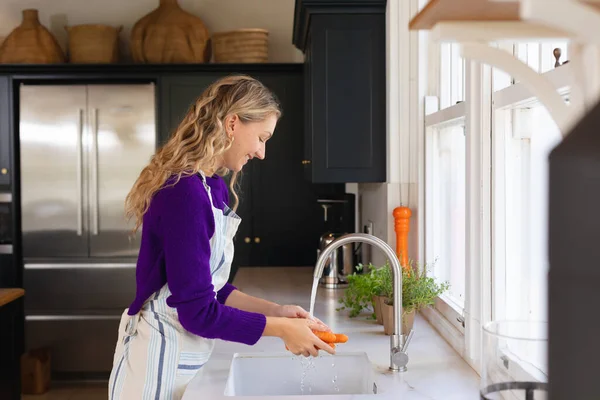 シンクでニンジンを洗うエプロンで台所に立つ笑顔の白人女性 家庭で一人で過ごす家庭生活 食べ物 健康的な食事 — ストック写真