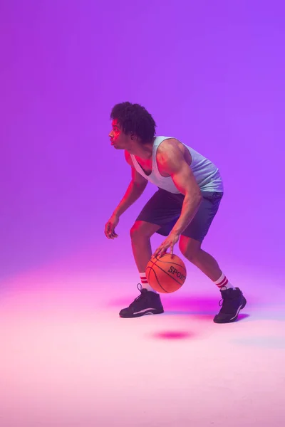 Image of biracial basketball player bouncing basketball on neon purple background. Sports and competition concept.