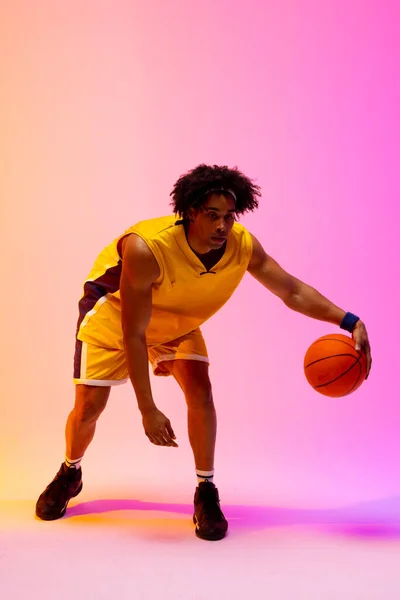 Image of biracial basketball player bouncing basketball on pink to orange background. Sports and competition concept.