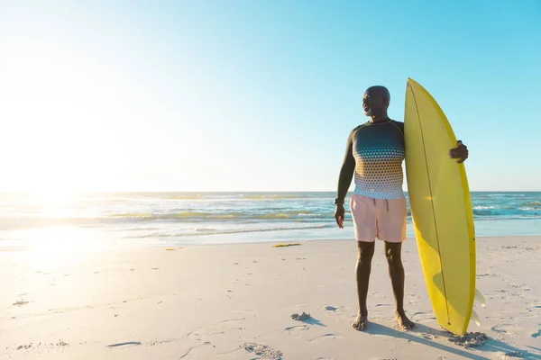 身穿黄色冲浪板的非洲裔美国老人站在沙滩上 面对着大海和晴朗的天空 复制空间 水上运动 — 图库照片
