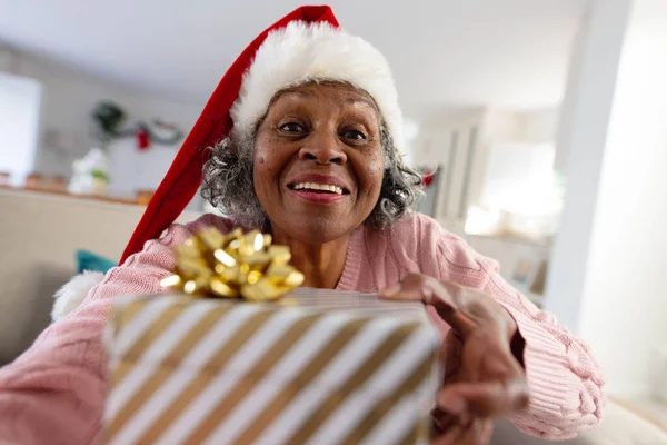 快乐的资深非洲裔美国女人戴着圣塔克洛斯帽 还带着礼物 独自呆在家里 利用技术 — 图库照片