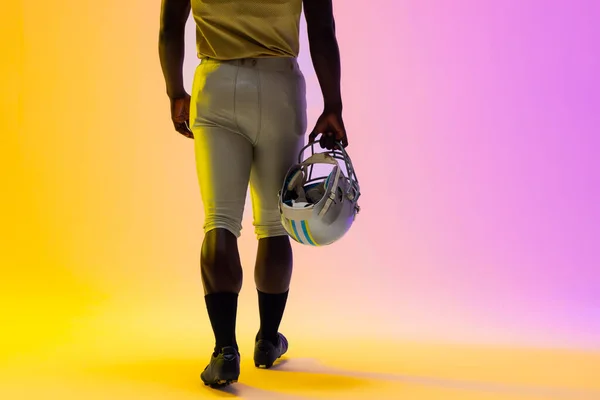 Midsection of african american male american football player with neon purple and yellow lighting. Sport, movement, training and active lifestyle concept.