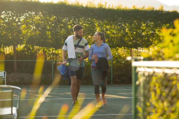 テニスコートでラケットを持って歩く幸せな白人夫婦のイメージ 活動的な品質の時間を一緒に過ごす — ストック写真