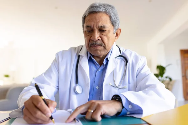 Starszy Dwurasowy Lekarz Siedzący Przy Biurku Robiący Notatki Podczas Konsultacji — Zdjęcie stockowe