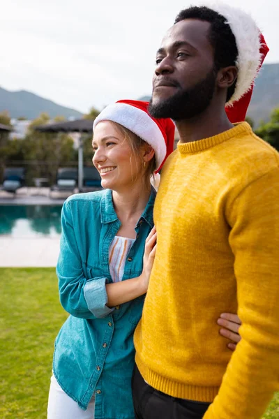 サンタの帽子で屋外に立ってクリスマスを祝う幸せな多様なカップルの垂直画像 クリスマス お祝い 包括性とライフスタイルの概念 — ストック写真
