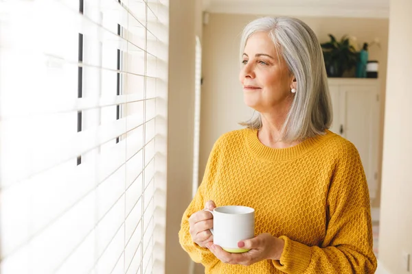 Przemyślana Biała Starsza Kobieta Stojąca Uśmiechem Przez Okno Domu Kawą — Zdjęcie stockowe