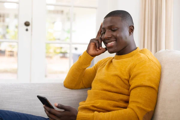 Leende Afrikansk Amerikansk Man Sitter Soffan Hemma Med Hjälp Smartphone — Stockfoto