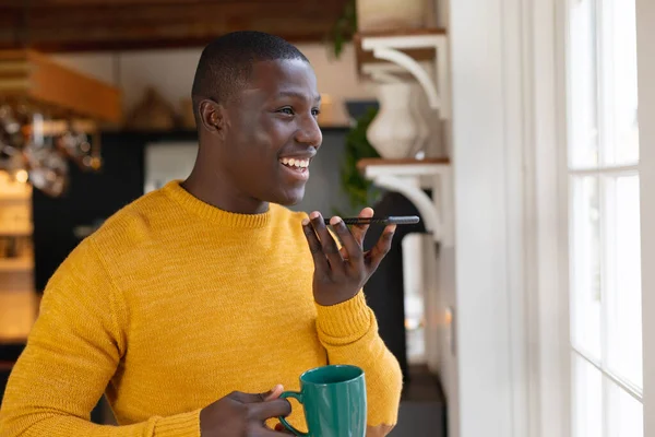 アフリカ系アメリカ人の男性がキッチンの窓から外を眺めながらコーヒーを飲みながらスマホで話しているのを笑っている 包括性 国内生活 積極性 家で一人で時間を過ごす — ストック写真