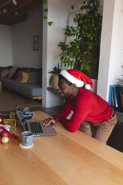 快乐的非洲裔美国人戴着桑塔帽 使用笔记本电脑 圣诞节时一个人呆在家里 — 图库照片