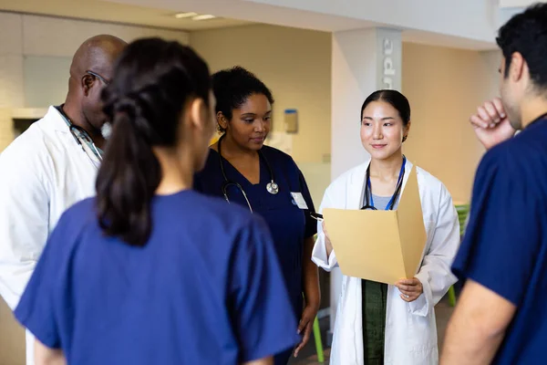 病院の廊下で多様な同僚と話すフォルダを持つ笑顔アジアの女性医師 医療サービス — ストック写真