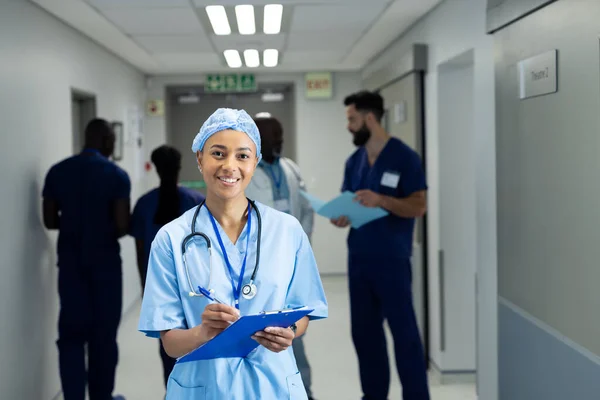 Portret Uśmiechniętej Dwurasowej Lekarki Piszącej Notatki Zatłoczonym Szpitalnym Korytarzu Przestrzeń — Zdjęcie stockowe