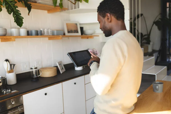 幸せなアフリカ系アメリカ人の男性がカウンターにもたれて台所でタブレットを使用しています 自宅だけで質の高い時間を過ごす — ストック写真