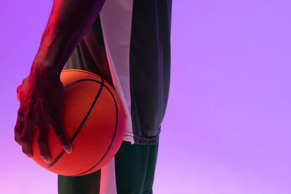 Image of midsection of african american basketball player with basketball on neon purple background. Sports and competition concept.