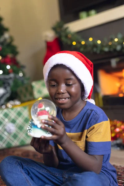 クリスマスの雪玉を保持し 次の暖炉に座っているアフリカ系アメリカ人の少年の垂直写真 クリスマス 子供時代 お祝いのコンセプト — ストック写真