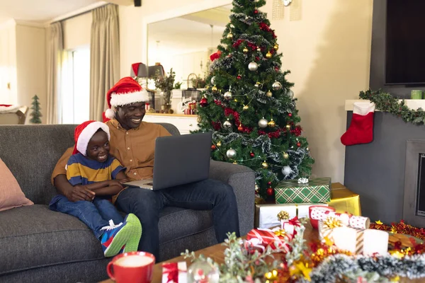 ソファに座ってノートパソコンを使っているアフリカ系アメリカ人の父親と息子 クリスマス 家族の時間と技術の概念を使用してお祝い — ストック写真