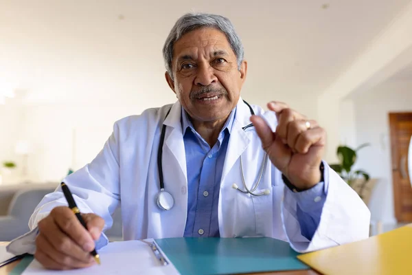 Starszy Dwurasowy Lekarz Siedzący Przy Biurku Rozmawiający Podczas Konsultacji Wideo — Zdjęcie stockowe