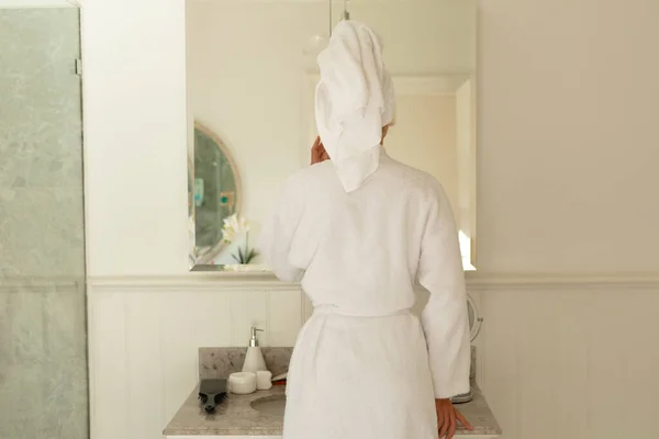 快乐的高加索女人穿着长袍 在镜子里看着自己的倒影 在浴室里微笑 独自在家度过美好时光 — 图库照片
