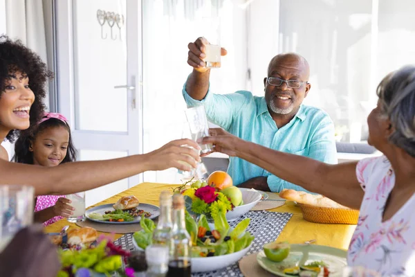 ダイニングテーブルで多世代家族とランチをしながら乾杯する多人種シニア男性 飲み物 変わらない 子供時代と退職の概念 — ストック写真