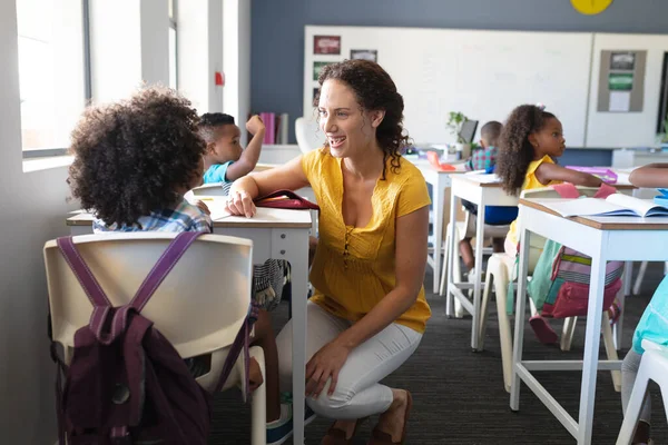 笑顔の白人の若い女性教師アフリカ系アメリカ人の少年と教室の机の上に座って話をする 変化のない教育子供時代教育学習学校の概念 — ストック写真