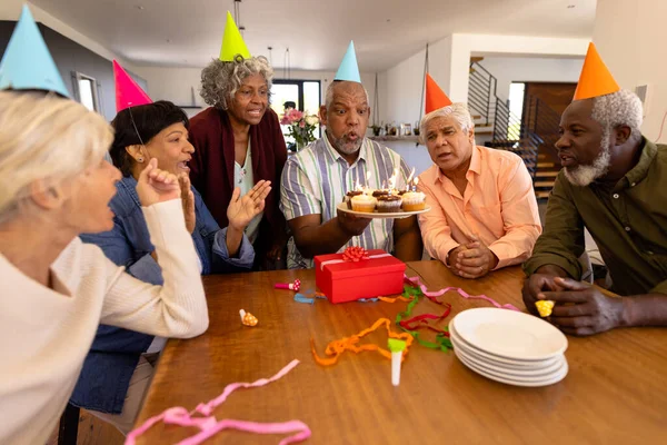 多种族的老朋友戴着派对帽 看着男人在养老院里吹蜡烛 辅助生活和退休 — 图库照片