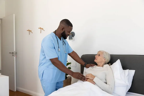 アフリカ系アメリカ人男性の健康労働者は 白人の高齢女性を支援するために自宅のベッドに横たわっている 医療と定年退職シニアライフスタイルの概念 — ストック写真