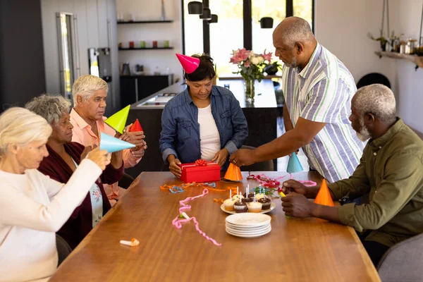 多种族老年人头戴派对帽 而妇女则在养老院的桌上打开生日礼物 辅助生活 — 图库照片