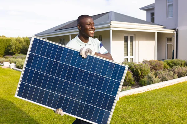 笑着非洲裔美国中年男子背着太阳能电池板在院子里与房子相对 不变的太阳能 绿色技术和可持续性概念 — 图库照片