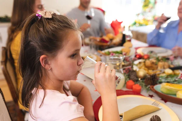 白人の少女の夕食のためのテーブルに座って ガラスを保持し 背景に彼女の家族と 家族の時間クリスマスのお祝い — ストック写真