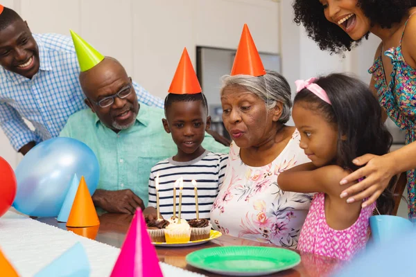 在与多种族家庭庆祝生日的同时 戴党的帽子的老年妇女在吹蜡烛 多代人 纸杯蛋糕 在一起 甜蜜的食物 — 图库照片
